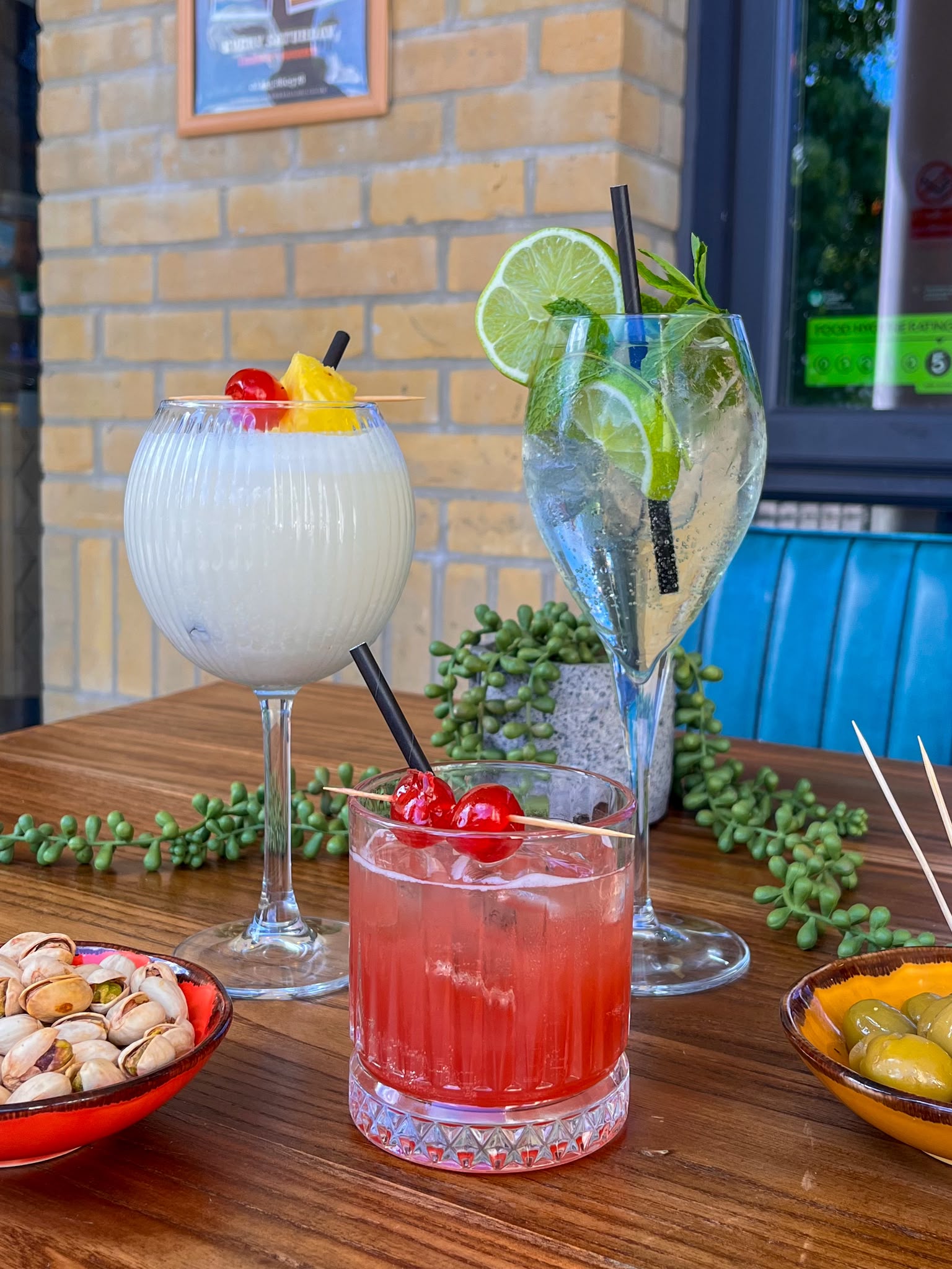 A piña colada, Hugo spritz and cherry amaretto sour on a table with bowls of pistachios and olives