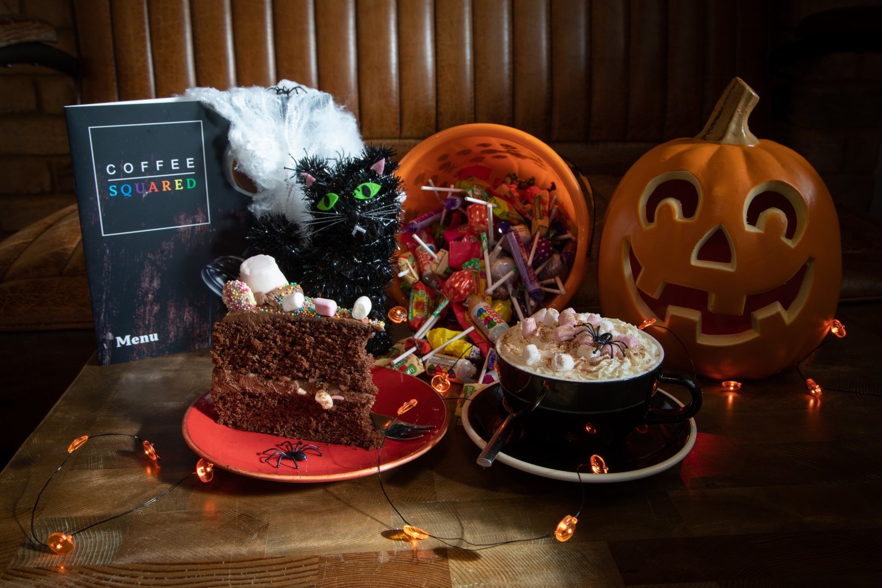 A Halloween table at Coffee Squared: carved pumpkin, chocolate cake, a bucket of sweets and a hot chocolate under orange fairy lights