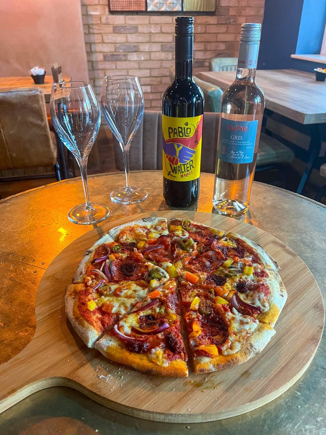 A freshly made pizza on a wooden board with red and rosé wine and two glasses